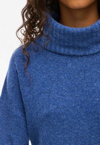 Kobaltblå turtleneck-tröja av strukturerat stickat material, med hög krage och lös passform. Lockigt hår synligt på ena sidan.