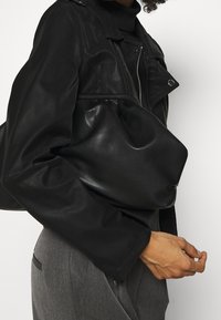 Giacca da biker in pelle nera con dettagli in zip argentati, indossata su un dolcevita nero, abbinata a una borsa nera con texture, pantaloni grigi.