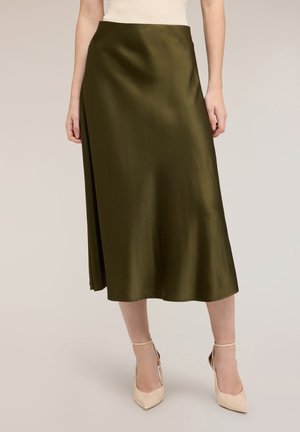 Vrouw draagt een olijfgroene satijnen midi-rok, gecombineerd met beige enkellaarzen met bandje, staand tegen een effen lichte achtergrond.