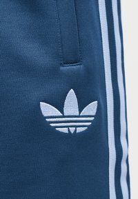 Μπλε ύφασμα με κεντημένο το λογότυπο Adidas trefoil σε λευκό και λευκές κάθετες ρίγες κατά μήκος της πλαϊνής ραφής.