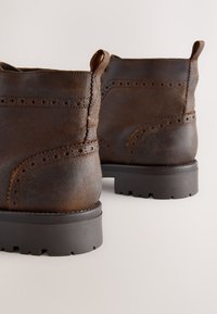 Braune Leder-Stiefeletten mit strukturiertem Finish, dekorativen Perforationen und schwarzer Gummi-Sohle. Mit Zuglaschen für einfaches An- und Ausziehen.