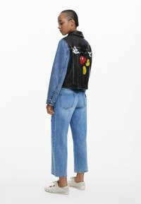 Denim jacket featuring a black leather back with a colorful cartoon graphic, paired with light blue denim jeans and white sneakers.
