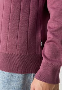 Pleten pulover v burgundski barvi z vertikalnim rebrastim vzorcem, strukturiran trup in prilegajoči se manšeti. Vidna stranska oznaka iz tkanine.