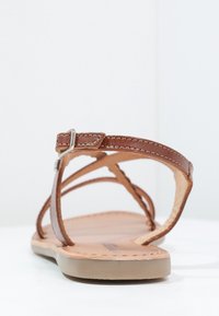 Hnědé kožené ploché sandály s tenkými popruhy a stříbrnou přezkou, zobrazené zezadu na bílé ploše a pozadí.