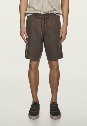 Mann trägt braune knielange Shorts mit Kordelzug in der Taille und schwarze Sneakers, steht vor einem schlichten hellen Hintergrund.