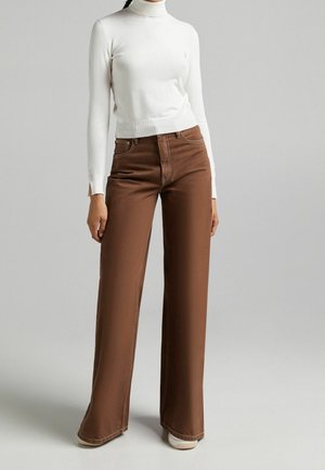 Woman wearing a white long-sleeve turtleneck and wide-leg brown pants standing against a gray background.