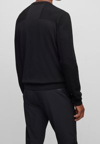 Svart stickad tröja med texturerade axelpaneler och ribbade mudd, matchad med mörka byxor som har en subtil varumärkeslogotyp.