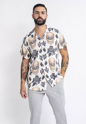 Camisa de manga corta con un patrón de calaveras y rosas, principalmente blanca con acentos en beige, gris y azul. Tejido suave, corte casual.