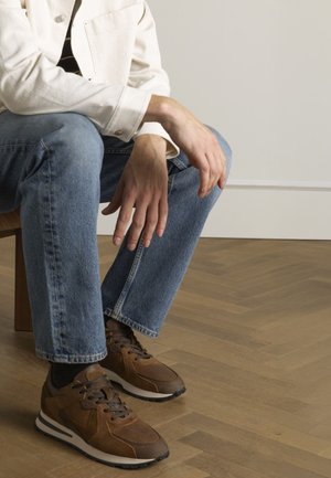Bruine leren sneakers met donkere veters en witte zolen, gedragen met lichtblauwe spijkerbroeken en een crèmekleurig overhemd met knopen.