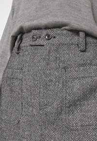 Grau gestreifte Hosen im Fischgrätmuster mit zwei schwarzen Knopfverschlüssen, seitlichen Taschen und einem strukturierten Stoff, der kleine rote und blaue Fasern aufweist.
