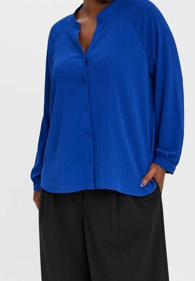 Personne portant une chemise bleue ample boutonnée à manches longues et un pantalon noir à jambes larges, main dans la poche.