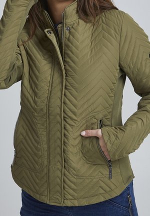 Femme portant une veste matelassée vert olive avec des poches zippées et des manches avec ouvertures pour le pouce, main dans la poche, et fermeture éclair partiellement fermée à l'avant.