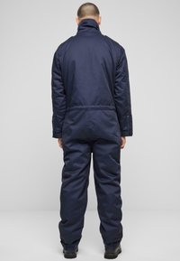 Tumši zilas krāsas izolēts kombinezons ar augstu apkakli, regulējamiem apkaklītes galiem un ar rāvējslēdzēju aizveramajiem kabatām. Gluda tekstūra ar brīvu piegriezumu.