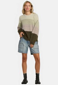 Maglione lavorato a maglia a righe verdi, beige e marroni; vestibilità oversize. Abbinato a shorts di denim e stivaletti neri alla caviglia. Tessuto liscio e testurizzato.