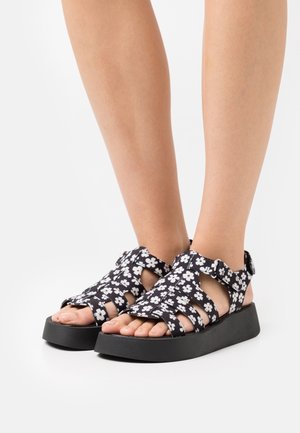Sandales à motif floral noir et blanc avec un design ouvert, une sangle ajustable et une épaisse semelle noire. Conviennent pour une tenue décontractée.