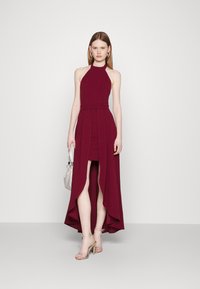 Burgundy halter neck dress with a fitted bodice and a high-low hemline, featuring a smooth texture and side slit. Worn with silver accessories.