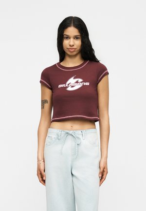 Ung kvinde med langt mørkt hår iført en vinrød Billabong crop top og lyseblå højttaljede bukser, stående mod en ensfarvet baggrund.