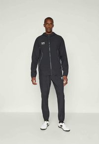 Hombre de pie vestido con una sudadera con cremallera negra de Under Armour, pantalones negros a juego y zapatillas blancas, frente a un fondo claro y sencillo.