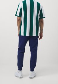 Zeleno-bíle pruhovaná bavlněná košile s krátkým rukávem spojená s tmavě modrými tepláky, obutá do bílých sportovních bot.