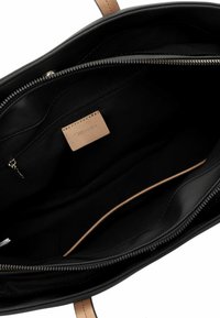 Intérieur d'un sac à main en cuir noir avec une texture lisse, une poche zippée et une étiquette en cuir beige. Présente une finition satinée et un design minimaliste.
