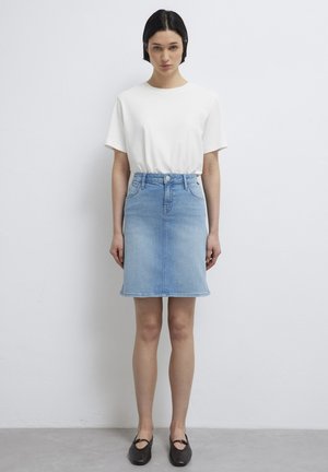 Frau steht vor einer schlichten weißen Wand, trägt ein weißes kurzärmeliges Hemd, einen hellblauen Jeansrock und schwarze flache Schuhe.