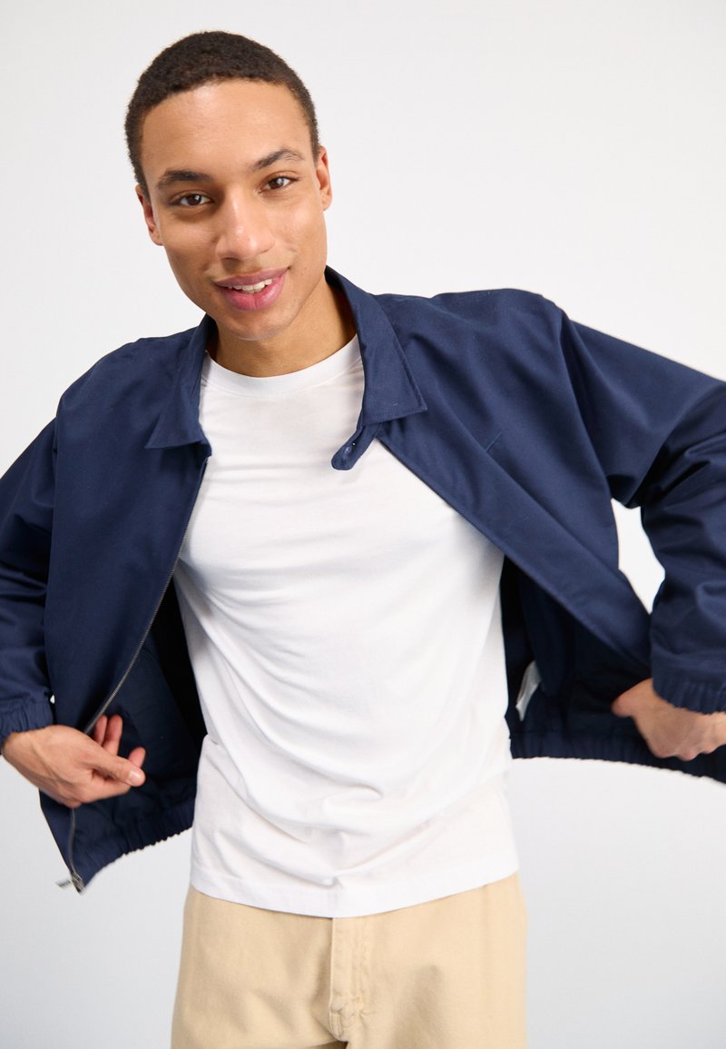 Veste légère bleu marine avec une fermeture éclair à l'avant, des poignets élastiques et un col, portée sur un t-shirt blanc et un pantalon beige.
