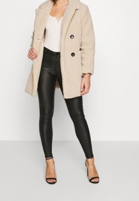 Beige teddyjacka med stora svarta knappar, parat med glänsande svarta läderleggings och svarta klacksandaler. Slät textur genom hela plagget.