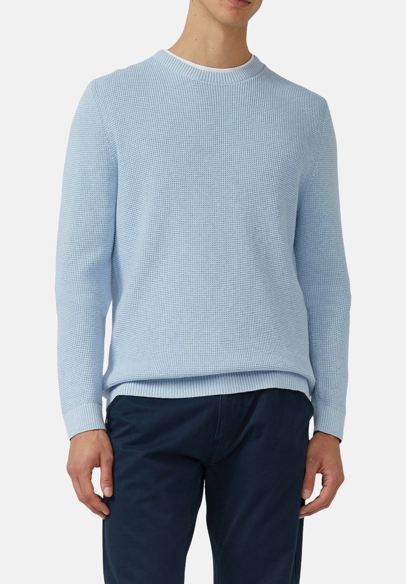 Mann in einem hellblauen strukturierten Pullover über einem weißen T-Shirt und dunklen Marinehosen, der vor einem einfarbigen Hintergrund steht.