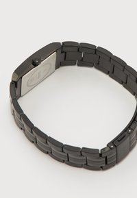 Μαύρο μεταλλικό λουράκι ρολογιού με ορθογώνια πίσω θήκη και κορώνα ρύθμισης σε φωτεινό φόντο, μερικώς ορατό κούμπωμα.