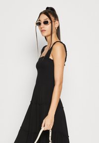 Robe noire à bretelles avec des détails volants, design à niveaux et corsage ajusté. Le mannequin porte des lunettes de soleil rectangulaires claires et tient un sac.