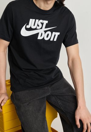 Osoba ubrana w czarną koszulkę Nike z napisem "Just Do It" i ciemne jeansy, siedząca na ułożonych stosach żółtych plastikowych skrzynek na jednolitym tle.