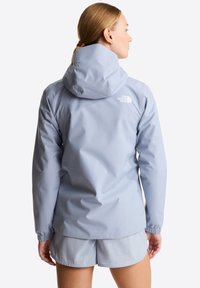 Γυναίκα με ανοιχτό μπλε αδιάβροχο με κουκούλα και ταιριαστά σορτς, εμφανίζεται από πίσω με φόντο απλό λευκό.