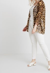 Transparante lange kimonojurk in bruin en oranje dierenprint over een wit topje, gecombineerd met witte strakke broek en witte sandalen met open teen.