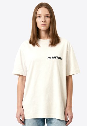 Jeune femme aux cheveux bruns et raides porte un t-shirt blanc ample avec le texte « YOU & ME TONIGHT » et un jean bleu clair, debout sur un fond blanc.