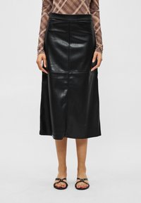 Melns ādas midi svārks, valkāts kopā ar melnām sandālēm ar atvērtām purngalām un garroku brūnu pleda topu, modelis redzams no vidukļa līdz kājām.