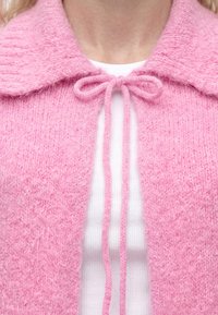 Gros plan sur un cardigan en maille rose avec un grand col noué en nœud papillon sur une chemise blanche côtelée.