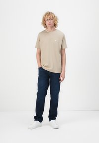 T-shirt beige à manches courtes avec un petit logo triangulaire, associé à un jean bleu foncé et des baskets blanches. Design simple et coupe classique.