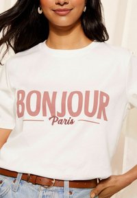 Bílý bavlněný tričko s potiskem "BONJOUR Paris" v růžové barvě. Má klasický kulatý výstřih a krátké rukávy. Kombinováno s hnědým pásem a světlými džíny.