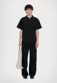 Svart polo-shirt med korte ermer og en treknappslukking, kombinert med vide svarte bukser. Holder en beige stoff totebag.