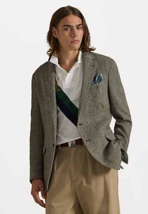 Blazer met een visgraatpatroon in grijs en beige, gedragen over een witte polo met een groene en marineblauwe streep, gecombineerd met beige broeken.