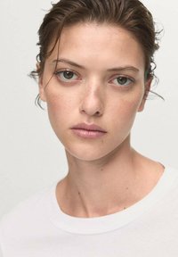 White t-shirt with a round neckline. Close-up of a person with short, wet hair and light skin, featuring green and brown eyes. Minimal background.