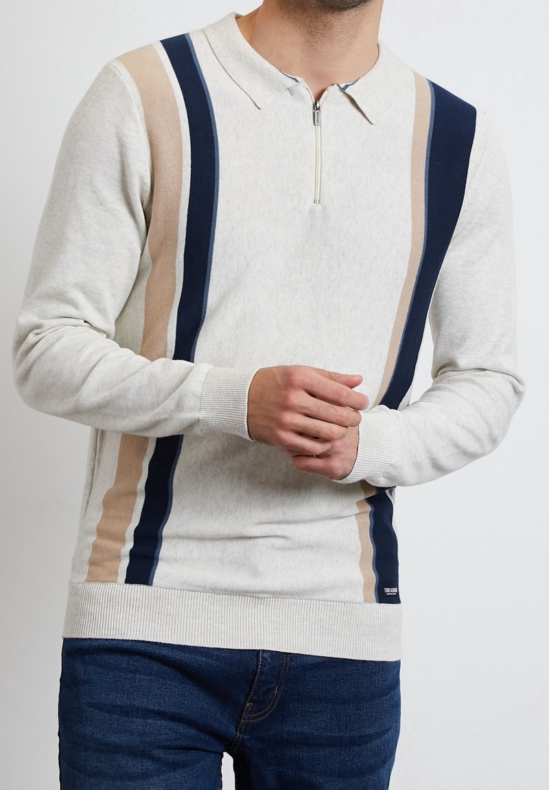 Homme portant un pull zippé gris clair avec des rayures verticales bleu marine et beige, associé à un jean bleu foncé.