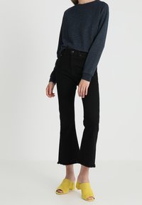 Zwarte uitlopende jeans met raw hem, gecombineerd met een navy gestructureerde trui en gele open teen muiltjes.