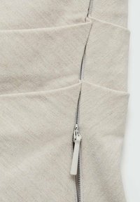 Gros plan d’un détail d’un vêtement en tissu beige avec une fermeture éclair blanche partiellement ouverte et des coutures visibles.