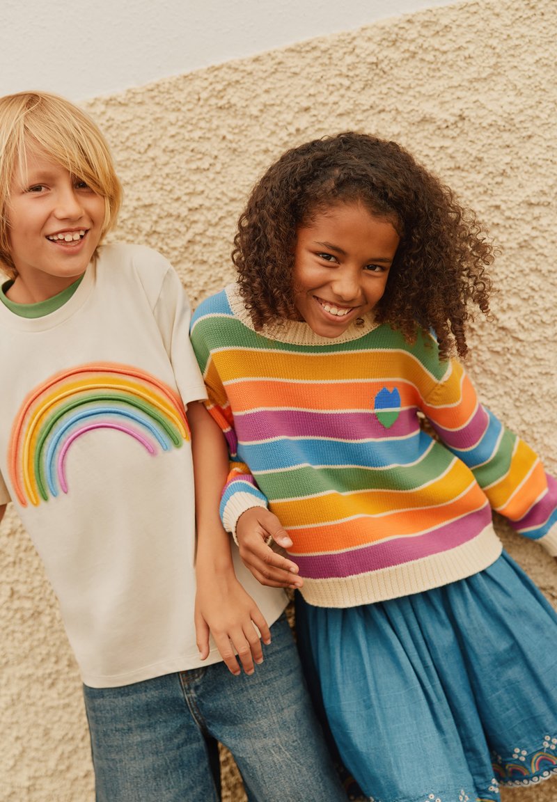 Dwoje dzieci stoi blisko siebie. Jedno z nich ma na sobie kremowy t-shirt z tęczowym wzorem. Drugie nosi sweter w wielokolorowe paski i niebieską spódniczkę.