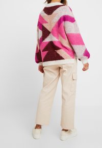 Maglioncino lavorato a maglia multicolore con motivi geometrici in rosa, marrone e crema. Abbinato a pantaloni cargo beige e scarpe di colore chiaro.