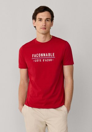 Hombre de pie con camiseta roja "Faconnable Côte d'Azur", pantalones beige y zapatillas negras, frente a un fondo gris liso.