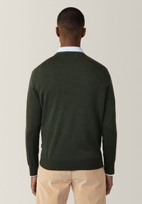 Maglione verde scuro con maniche lunghe e polsini a costine, dotato di scollo a girocollo e texture liscia. Indossato sopra una camicia azzurro chiaro.