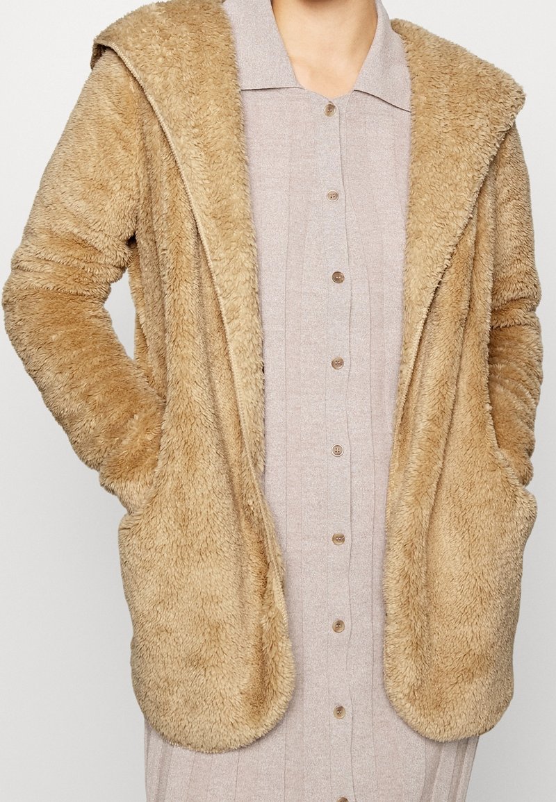Personne portant une robe beige boutonnée avec une veste à capuche duveteuse marron clair, mains dans les poches de la veste, montrée du cou à mi-cuisse.