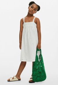 Vit ärmlös klänning med hålbroderi; har ett palm-mönsterdesign. En grön stickad tote-väskan med blommiga detaljer; guld sandaler.